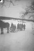 Ett sällskap färdas vintertid på en snötäckt landsväg med häst och släde. Några personer sitter i släden medan andra står bredvid på skidor. Öppet landskap med gårdsbebyggelse och telefonstolpar i bakgrunden.