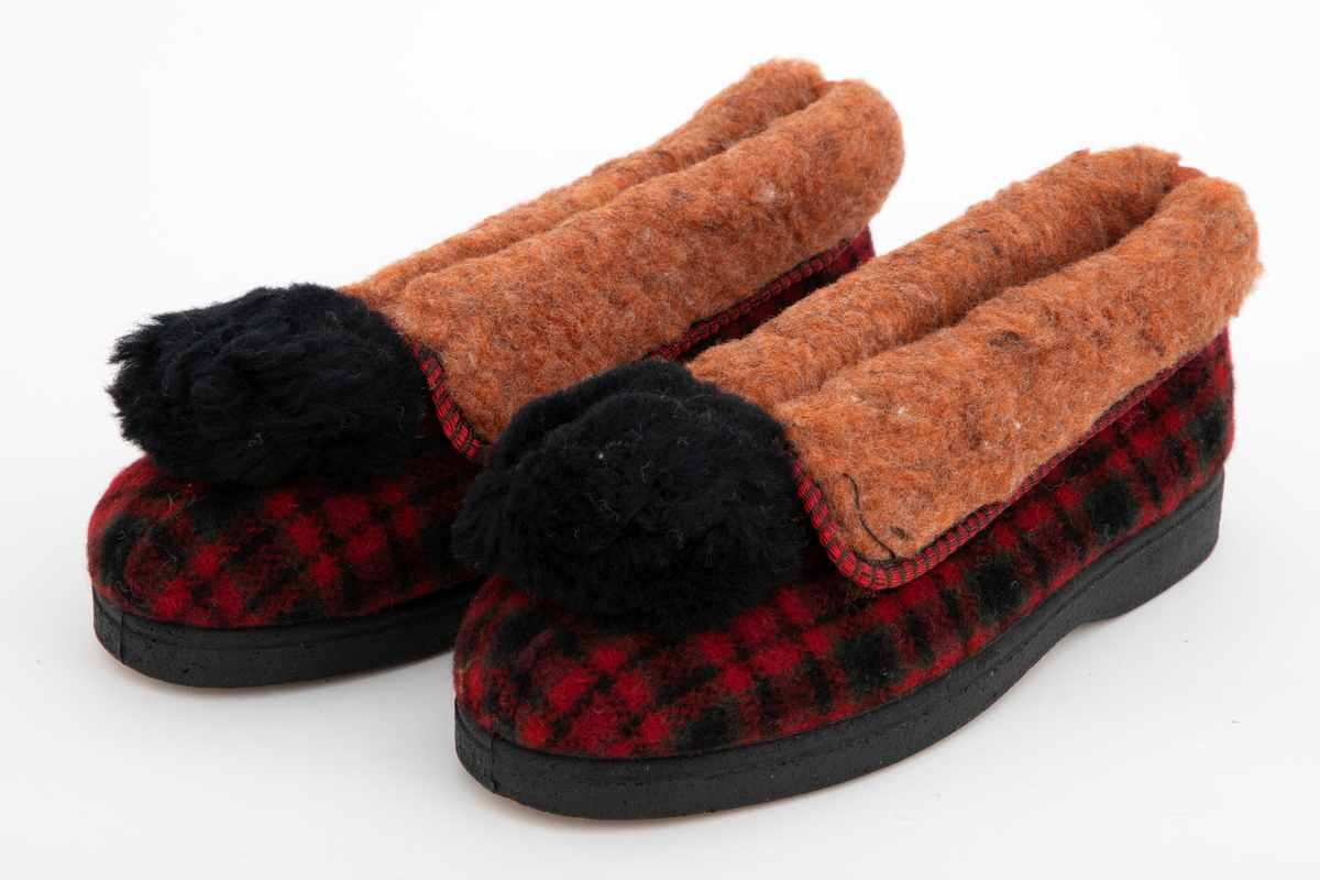 Tøfler i rutete ull i rødt, svart og grønt med nedbrettet og limt overkant i rødbrun, lodden viskosepels. Tøflene er innvendig fôret med samme materiale. Stor svart pompong på forstykket.