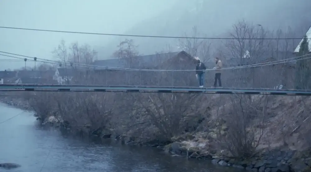 Bilde av hengebro med trær og hus i bakgrunnen i et tåkete landskap. To mennesker er på broen