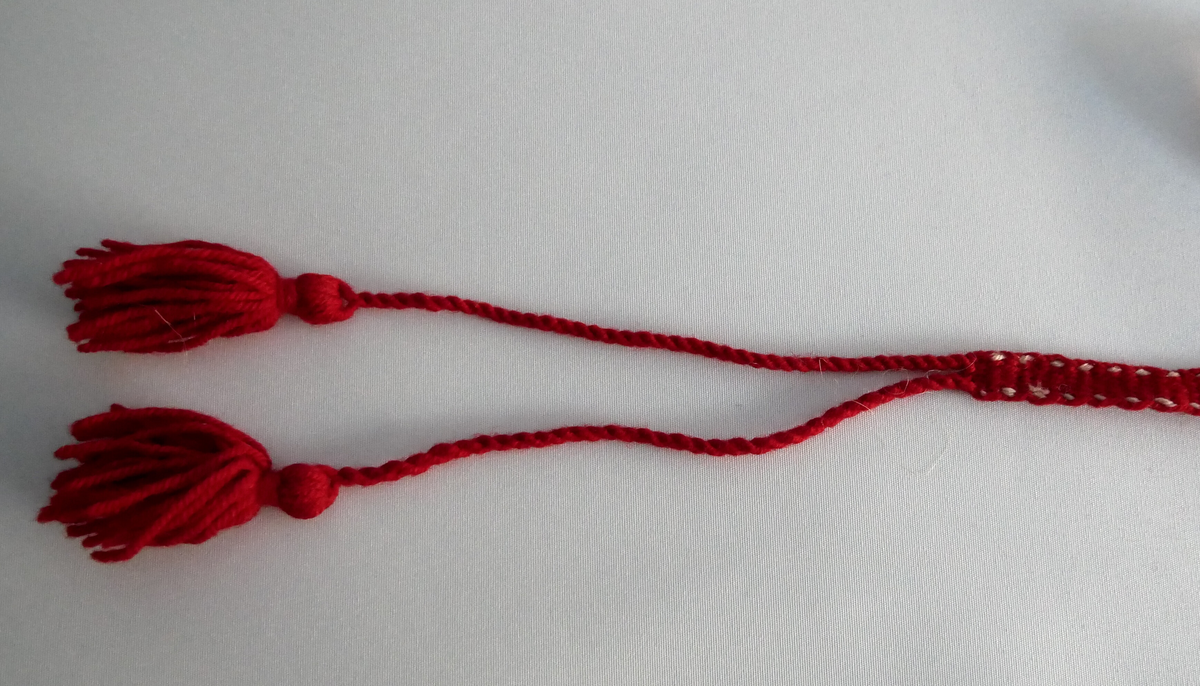 Grindvove band i raudt ullgarn med ein tråd i kvitt ullgarn på kvar side. I eit stykke på midten av bandet er det fletta saman to band. I kvar ende er det to duskar. 