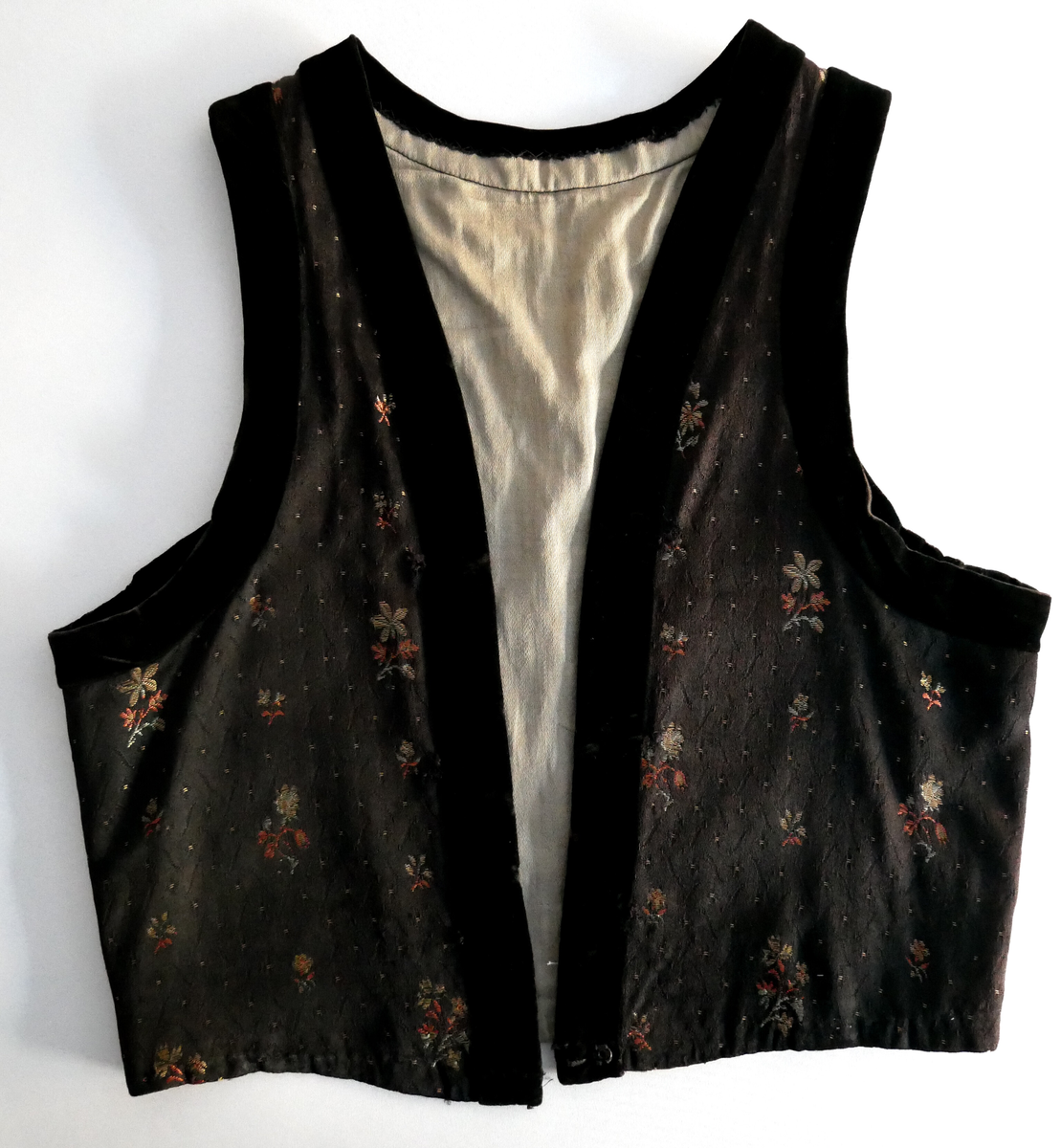 Livet er svart i botn med spetter og grøne og rosa blomar. Livet har v-forma opning framme og kring armhol og opning er det svarte fløyelsband. Liver er fora med beige bomullsstoff. 