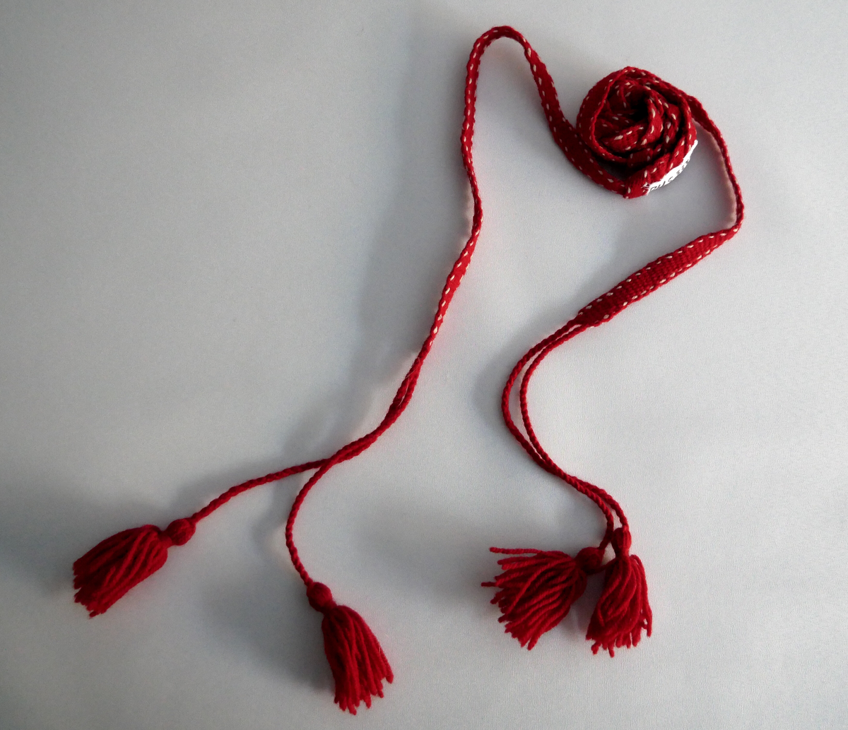 Grindvove band i raudt ullgarn med ein tråd i kvitt ullgarn på kvar side. I eit stykke på midten av bandet er det fletta saman to band. I kvar ende er det to duskar. 