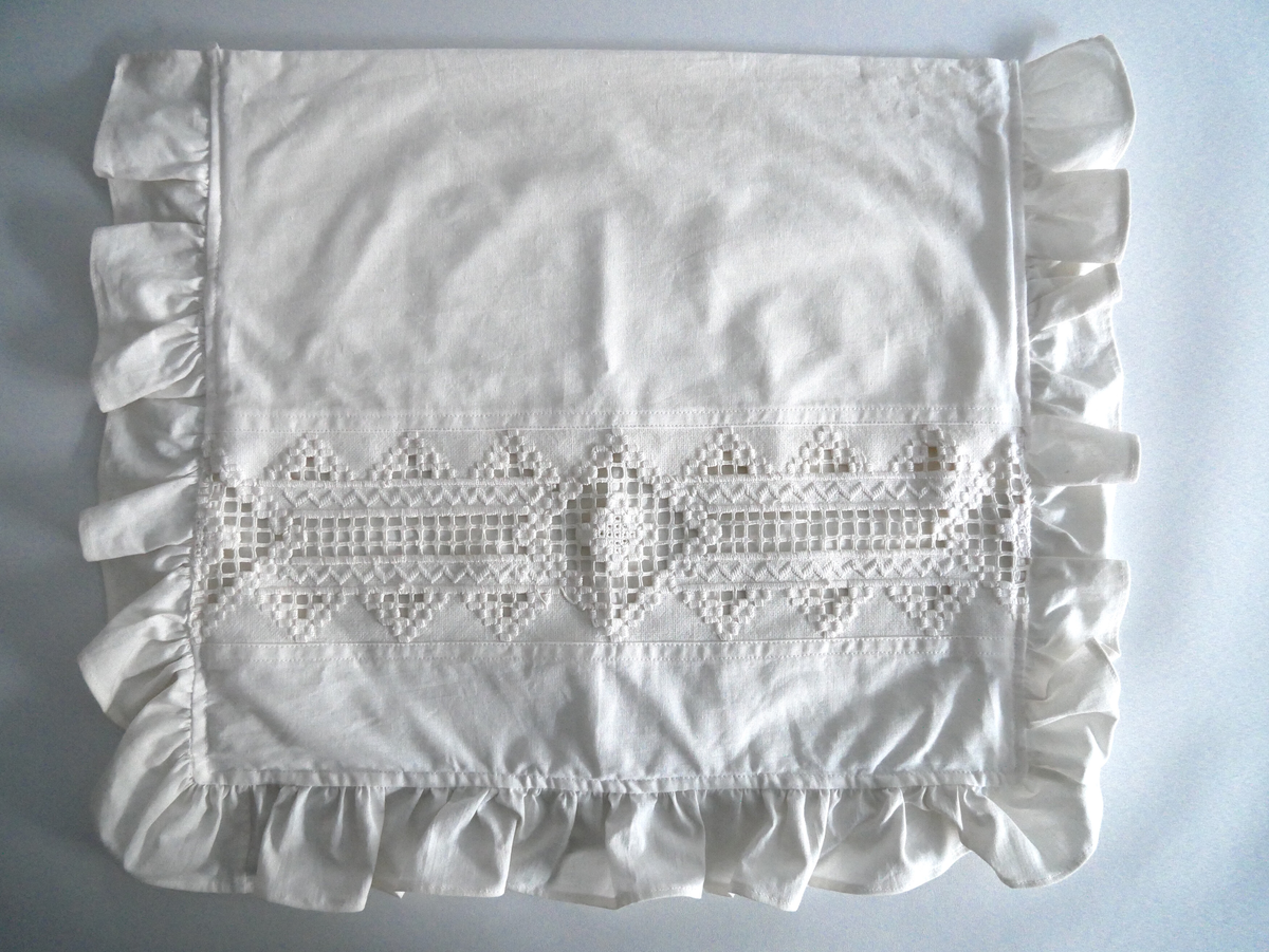 Kvitt putevar med rynkekappe rundt tre av kantane, med innfelt broderi i hardangersaum. Broderiet er 12, 0 cm høgt. Putevaret er laga som ei sengetøy-pute, men broderiet vil ikkje tole å bli brukt til å sove på. 