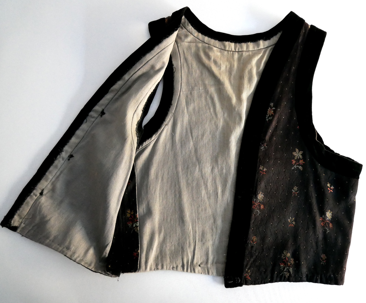 Livet er svart i botn med spetter og grøne og rosa blomar. Livet har v-forma opning framme og kring armhol og opning er det svarte fløyelsband. Liver er fora med beige bomullsstoff. 