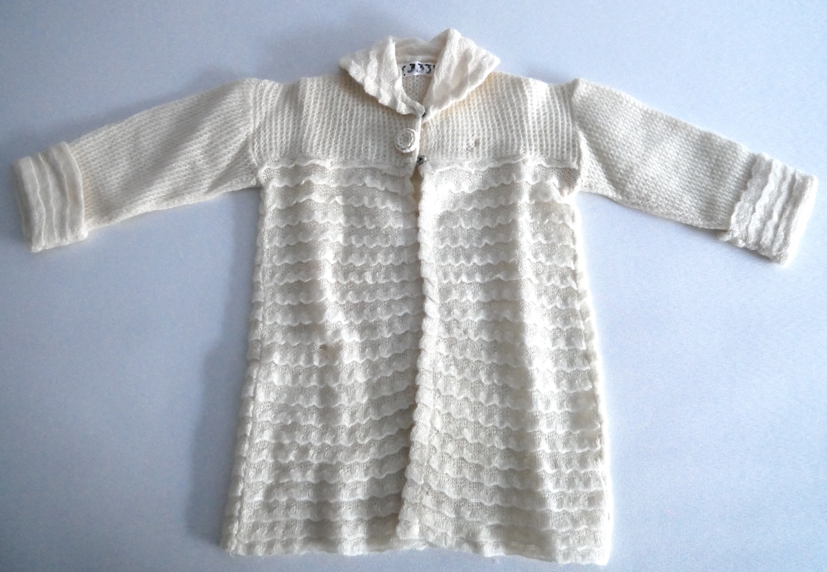 Jakke og lue i ubleika bomull,  industrielt framstilt i ulike strikketeknikkar. Jakka har krage og oppbrett på armane. Ho er åpen framme med to trykk-knappar og ein pynteknapp. Lua har syntetiske blonder rundt åpningen med band til knyting.  