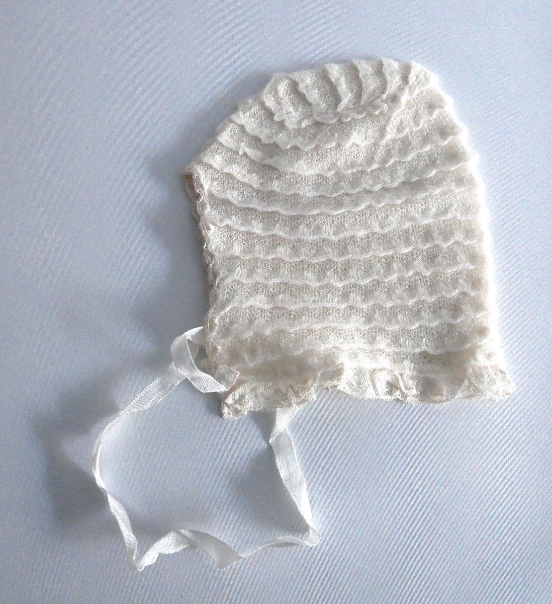 Jakke og lue i ubleika bomull,  industrielt framstilt i ulike strikketeknikkar. Jakka har krage og oppbrett på armane. Ho er åpen framme med to trykk-knappar og ein pynteknapp. Lua har syntetiske blonder rundt åpningen med band til knyting.  