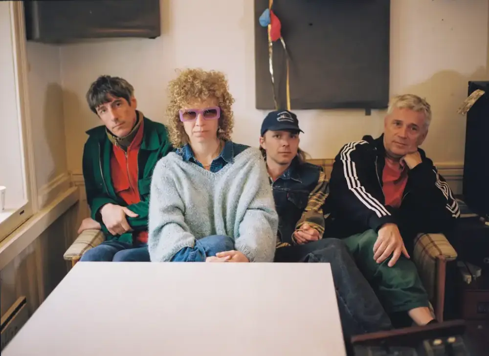 fire bandmedlemar sit ein gamal sofa bakom eit kvit firkanta bord