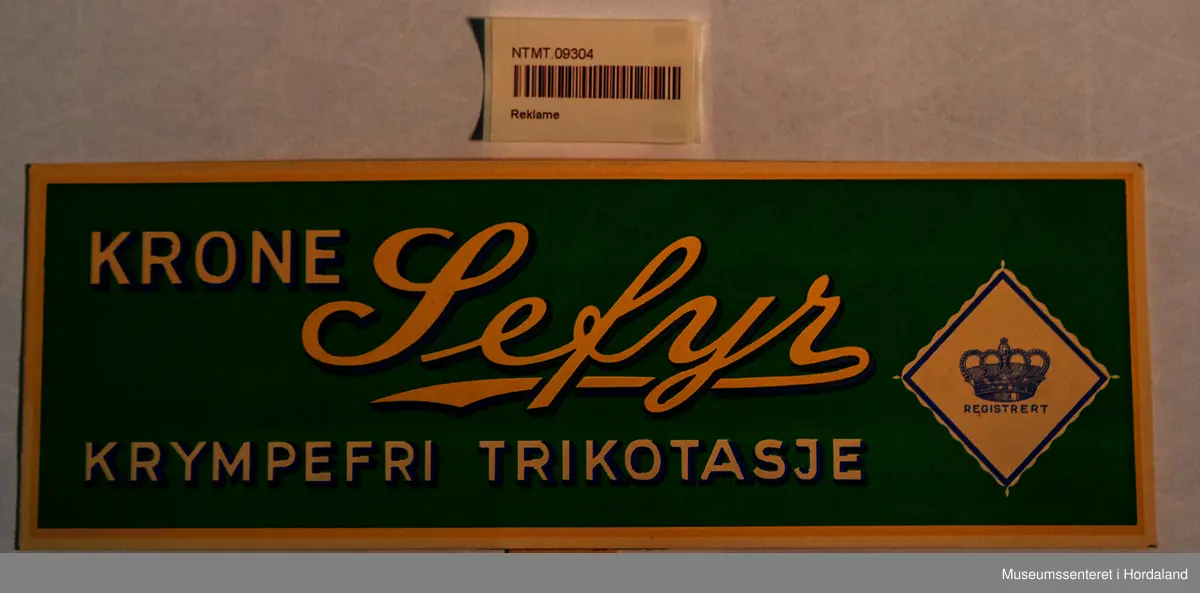 Reklameplakat på papp/kartong. Med "fot" på baksida slik at den kan stå på ein disk eller i ei hylle.