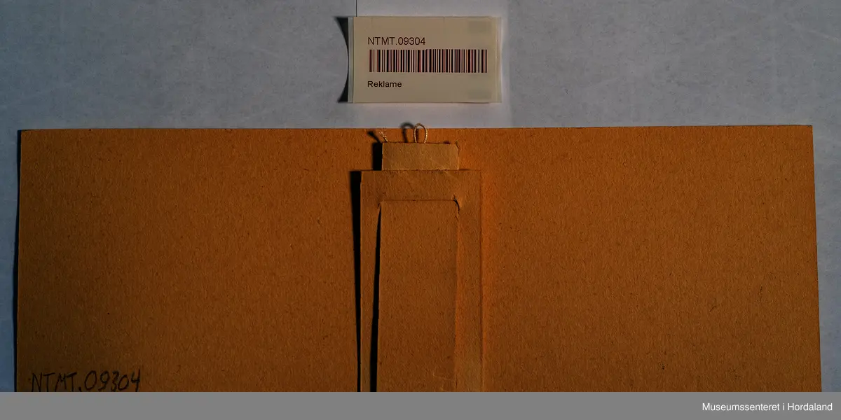 Reklameplakat på papp/kartong. Med "fot" på baksida slik at den kan stå på ein disk eller i ei hylle.