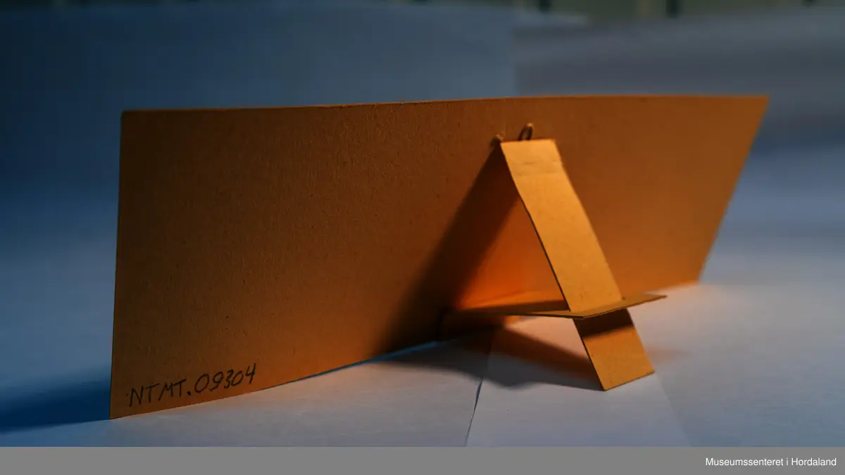 Reklameplakat på papp/kartong. Med "fot" på baksida slik at den kan stå på ein disk eller i ei hylle.