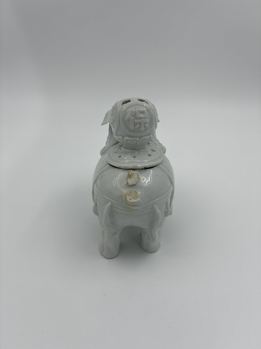 Prydnadsfigur i form av hund i kinesisk stil.
Figuren är ihålig och har på ryggen en öppning försedd med ett lock. 
Locket har en stor, delvis genombruten knopp prydd med fyra kinesiska skrivtecken i relief.
Vit glasyr.

Funktion: Möjligen rökelsekar