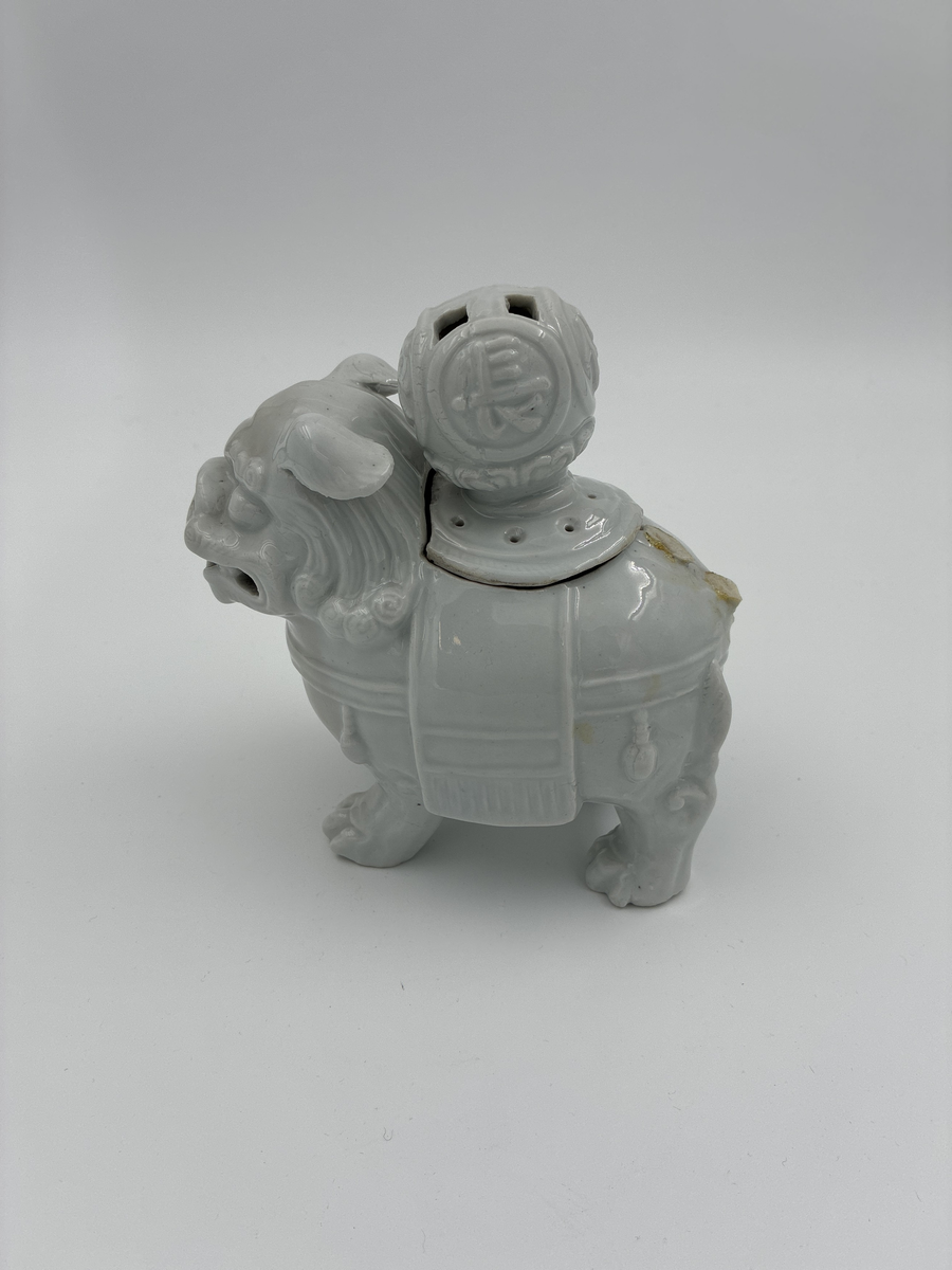 Prydnadsfigur i form av hund i kinesisk stil.
Figuren är ihålig och har på ryggen en öppning försedd med ett lock. 
Locket har en stor, delvis genombruten knopp prydd med fyra kinesiska skrivtecken i relief.
Vit glasyr.

Funktion: Möjligen rökelsekar