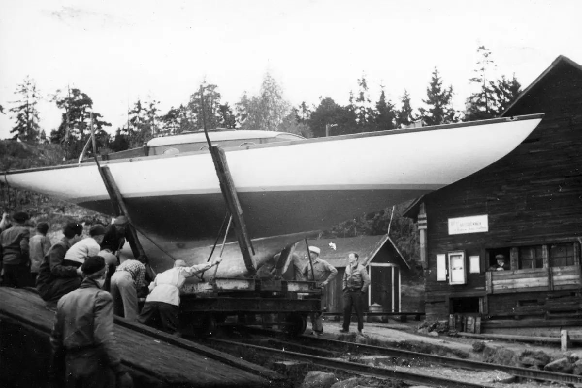 Sjösättningen av Fritiof Colbings egenbyggda segelbåt "Sigrun". En grupp män står runt båten, som precis blivit lyft till en en ställning som står på en liten räls. Vid den så kallade Saltladan, som låg vid de gamla lokstallarna i Nynäshamns hamns norra område. Båten byggdes åren 1948-1951. 