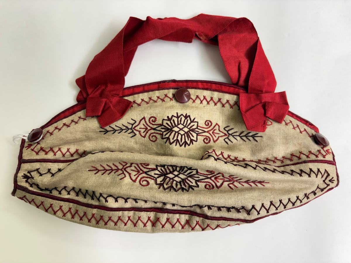 Liten väska för stickning. Bottentyg i beige linnetyg. 5 st ovala tygbitar hopsydda med passpoalerad rött linne mellan bitarna samt runt ytterkanten. Broderad med mönster, blommor, snirklar i rött och brunt med stjälkstygn och kråkspark. 3 st bruna knappar på ena långsidan andra en virkad trådögla övriga saknas. Fodrad med rött linnetyg. Handtag av rött sidenband avslutad med rosett.
Användes för att bära med sig liten stickning.