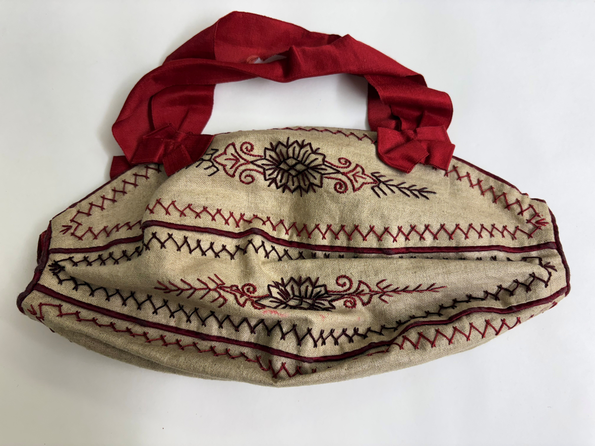 Liten väska för stickning. Bottentyg i beige linnetyg. 5 st ovala tygbitar hopsydda med passpoalerad rött linne mellan bitarna samt runt ytterkanten. Broderad med mönster, blommor, snirklar i rött och brunt med stjälkstygn och kråkspark. 3 st bruna knappar på ena långsidan andra en virkad trådögla övriga saknas. Fodrad med rött linnetyg. Handtag av rött sidenband avslutad med rosett.
Användes för att bära med sig liten stickning.