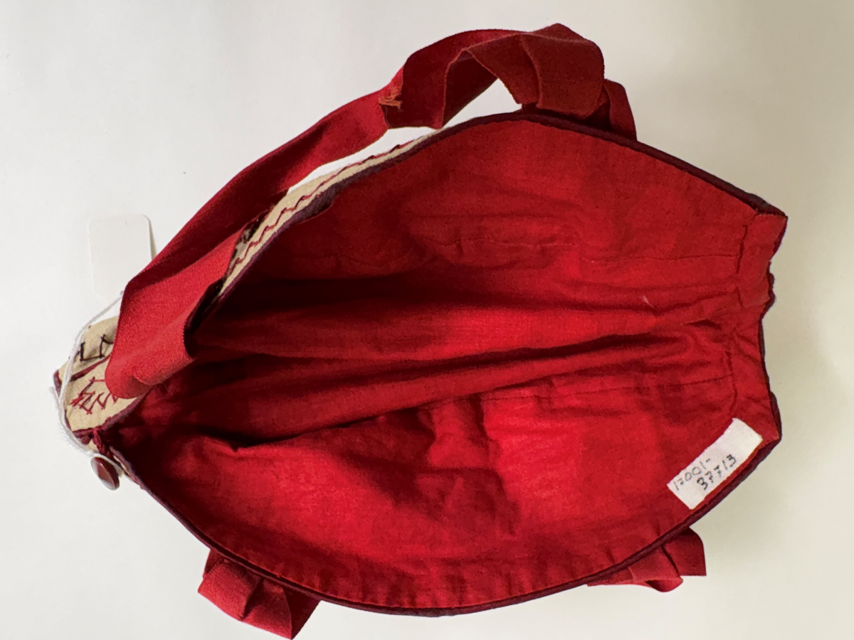 Liten väska för stickning. Bottentyg i beige linnetyg. 5 st ovala tygbitar hopsydda med passpoalerad rött linne mellan bitarna samt runt ytterkanten. Broderad med mönster, blommor, snirklar i rött och brunt med stjälkstygn och kråkspark. 3 st bruna knappar på ena långsidan andra en virkad trådögla övriga saknas. Fodrad med rött linnetyg. Handtag av rött sidenband avslutad med rosett.
Användes för att bära med sig liten stickning.