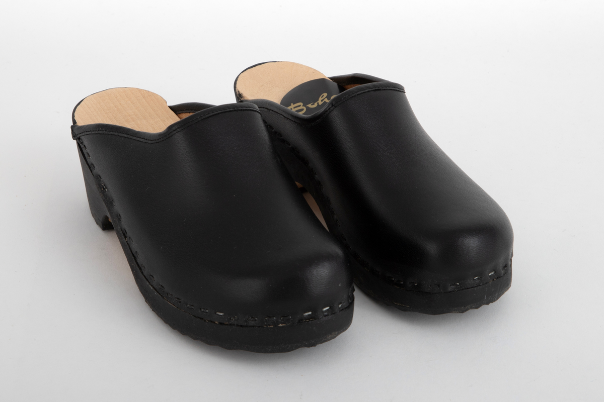 Tresko til barn i svart lær og svartmalt såle av tre. Læret er festet til sålen med stifter. Gummibelegg under sålen.