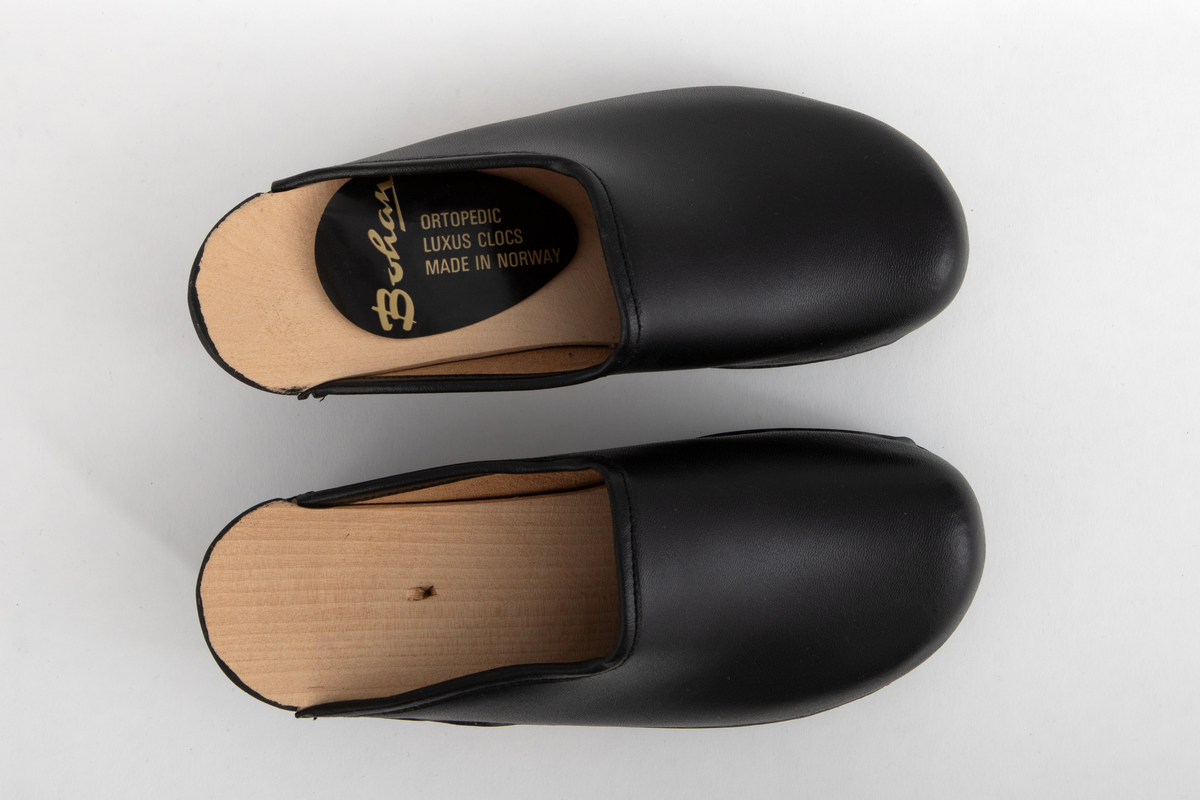 Tresko til barn i svart lær og svartmalt såle av tre. Læret er festet til sålen med stifter. Gummibelegg under sålen.