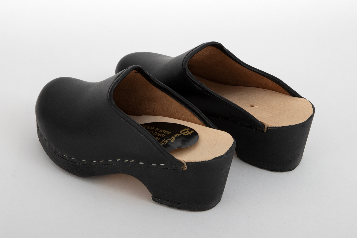 Tresko til barn i svart lær og svartmalt såle av tre. Læret er festet til sålen med stifter. Gummibelegg under sålen.