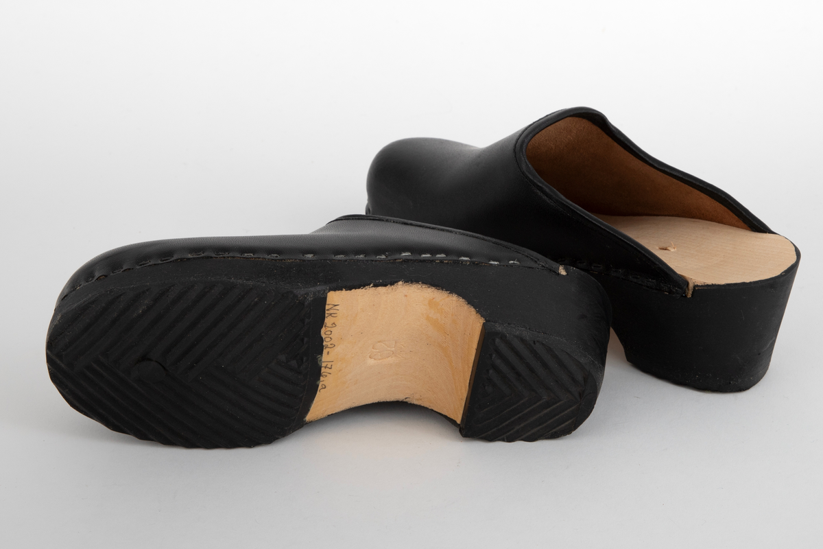 Tresko til barn i svart lær og svartmalt såle av tre. Læret er festet til sålen med stifter. Gummibelegg under sålen.