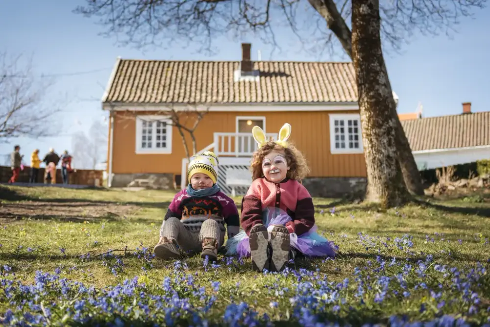 To barn sitter i hagen utenfor Munchs lille gule hus, med mange blå blomster på bakken foran dem. Det ene barnet er pyntet med kaninører og ansiktsmlaing.
