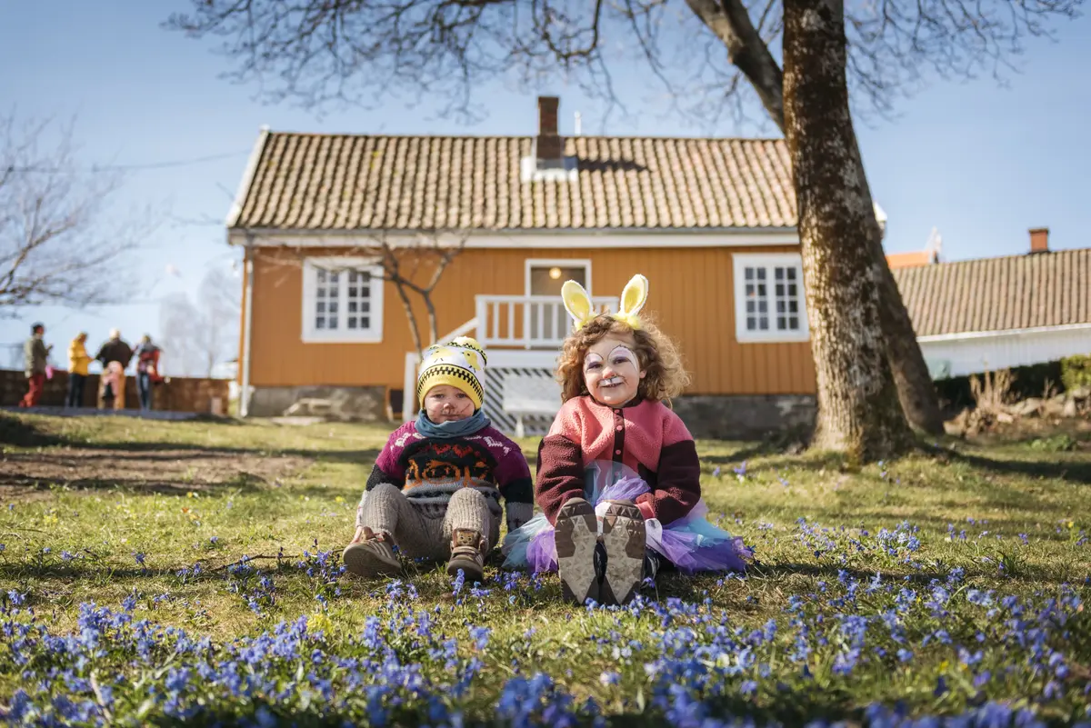 To barn sitter i hagen utenfor Munchs lille gule hus, med mange blå blomster på bakken foran dem. Det ene barnet er pyntet med kaninører og ansiktsmlaing.
