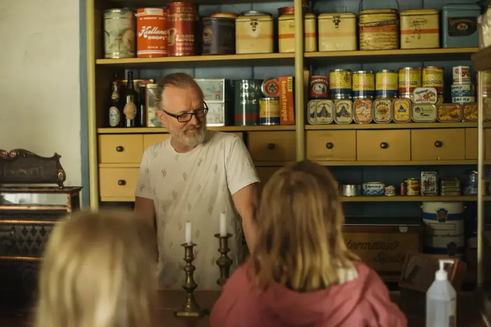 Erlend står bak disken med masse gamle hermetikkbokser, hovedsaklig gule, i hylla bak. Vi ser bakhodet til to barn foran disken.