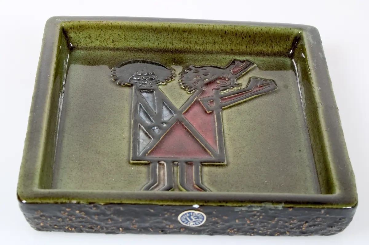 Fyrkantigt fat med kant med brunsvart glasyr och ojämn yta på utsidan och grönbrun glasyr på insidan. Reliefdekor i form av två stiliserade barn i rött, blått och svart. Text i relief: Folke Bernadottehemmet Uppsala Sweden, Hofmann, UE Sweden Abg. Tillverkarens pappersetikett.