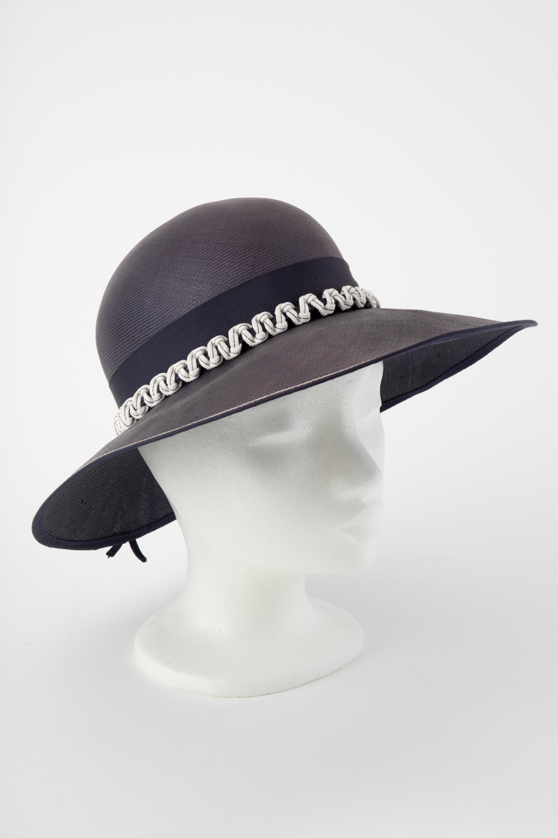 Midnattblå stråhatt av meget fint og tynt strå. Høy, rund pull, bred og hvelvet brem. Bredt hattebånd av tilsvarende blått ripsbånd, dvs to-skafts bånd med innslagseffekt. Svettebånd av smalt ripsbånd i samme farge. Hatten er i tillegg prydet utenpå nedre halvdel av hattebåndet med et kraftig, hvitt bånd med blå stikning, knytt i sikksakk-mønster omkring et blått bånd. Bremmen er noe bredere foran enn midt bak, og kantet med et smalt, blått bånd med hvit stikning. En sløyfelignende båndløkke av smalt ripsbånd. er festet på hattebåndet midt bak og ligger ut over bremmen. Båndet er gjort smalt ved at det er brettet dobbelt og holdt sammen av en maskinsøm. Ufôret. Ingen etikett.