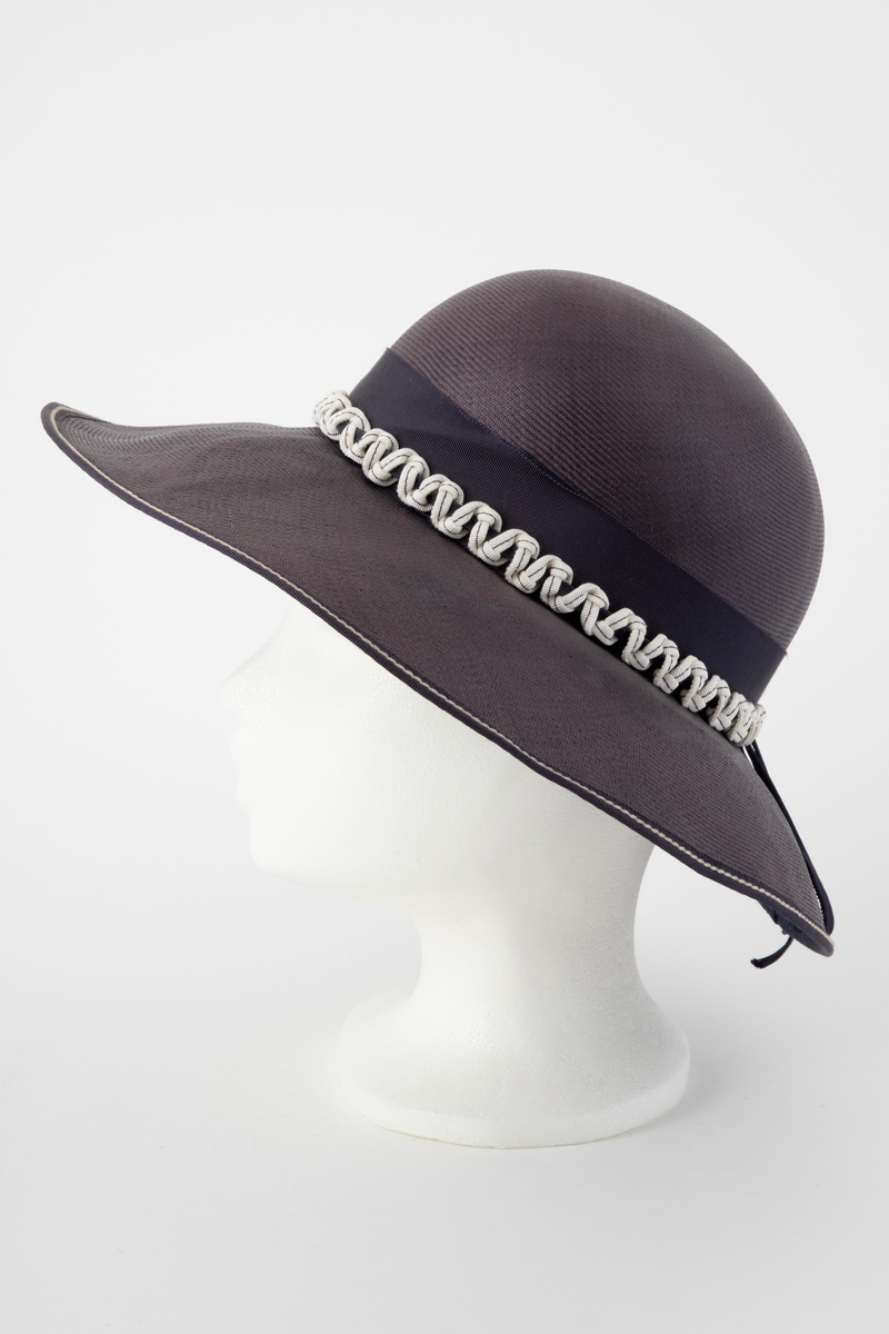 Midnattblå stråhatt av meget fint og tynt strå. Høy, rund pull, bred og hvelvet brem. Bredt hattebånd av tilsvarende blått ripsbånd, dvs to-skafts bånd med innslagseffekt. Svettebånd av smalt ripsbånd i samme farge. Hatten er i tillegg prydet utenpå nedre halvdel av hattebåndet med et kraftig, hvitt bånd med blå stikning, knytt i sikksakk-mønster omkring et blått bånd. Bremmen er noe bredere foran enn midt bak, og kantet med et smalt, blått bånd med hvit stikning. En sløyfelignende båndløkke av smalt ripsbånd. er festet på hattebåndet midt bak og ligger ut over bremmen. Båndet er gjort smalt ved at det er brettet dobbelt og holdt sammen av en maskinsøm. Ufôret. Ingen etikett.