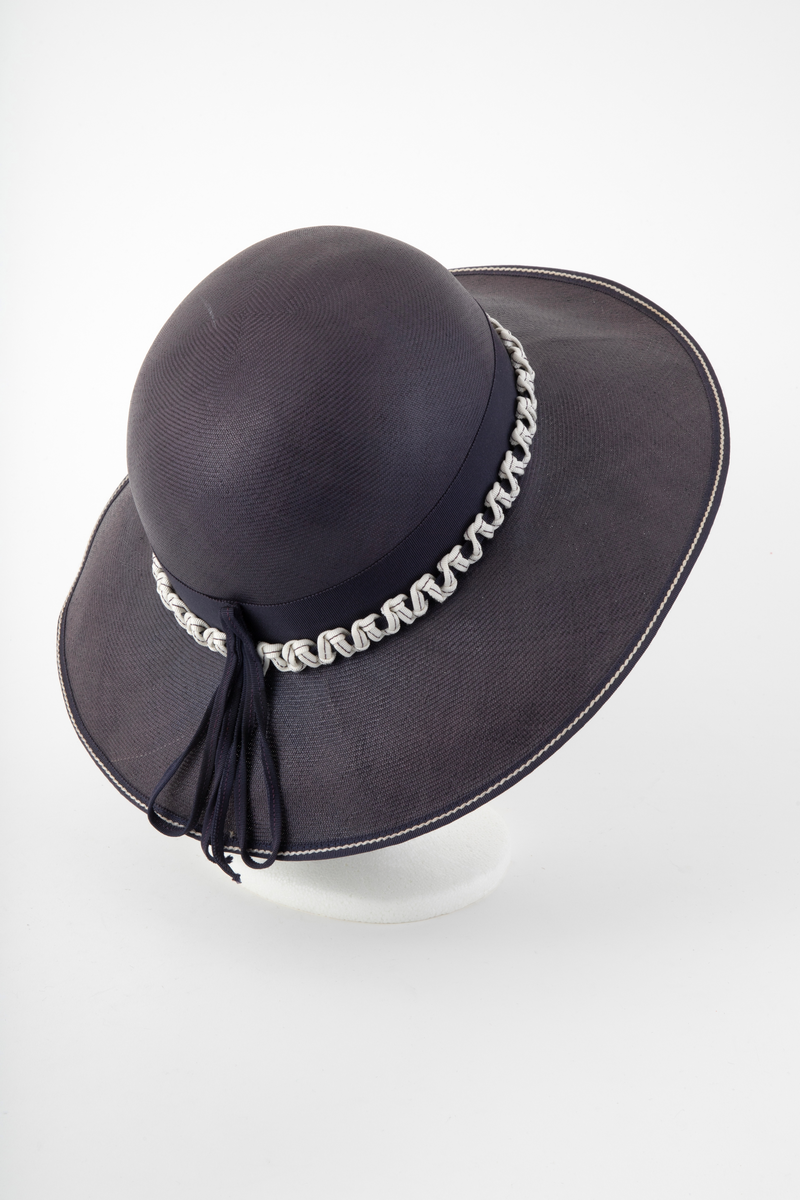 Midnattblå stråhatt av meget fint og tynt strå. Høy, rund pull, bred og hvelvet brem. Bredt hattebånd av tilsvarende blått ripsbånd, dvs to-skafts bånd med innslagseffekt. Svettebånd av smalt ripsbånd i samme farge. Hatten er i tillegg prydet utenpå nedre halvdel av hattebåndet med et kraftig, hvitt bånd med blå stikning, knytt i sikksakk-mønster omkring et blått bånd. Bremmen er noe bredere foran enn midt bak, og kantet med et smalt, blått bånd med hvit stikning. En sløyfelignende båndløkke av smalt ripsbånd. er festet på hattebåndet midt bak og ligger ut over bremmen. Båndet er gjort smalt ved at det er brettet dobbelt og holdt sammen av en maskinsøm. Ufôret. Ingen etikett.