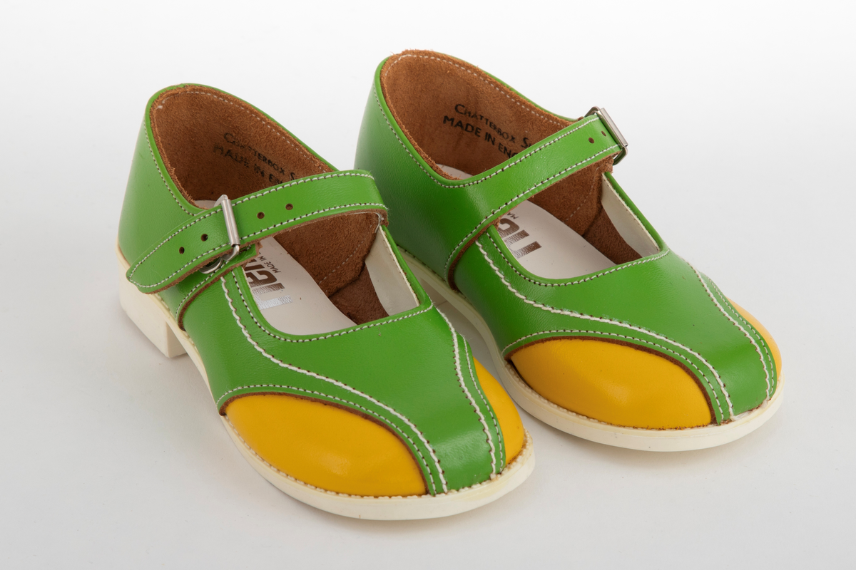 Barnesko i gressgrønt lær med gule detaljer på hver side av forstykket. Hvite stikninger og vristrem med spenne. Skoene har hvite såler i syntetisk gummi.