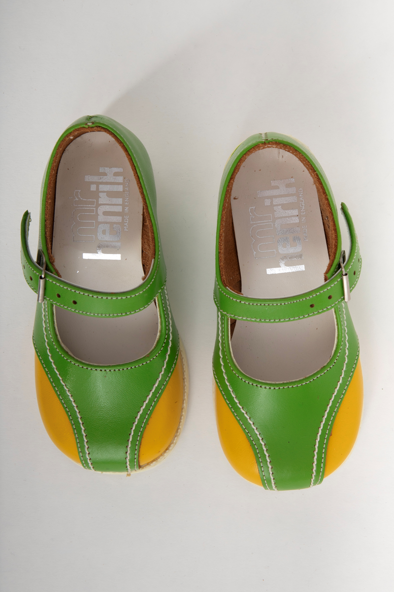 Barnesko i gressgrønt lær med gule detaljer på hver side av forstykket. Hvite stikninger og vristrem med spenne. Skoene har hvite såler i syntetisk gummi.