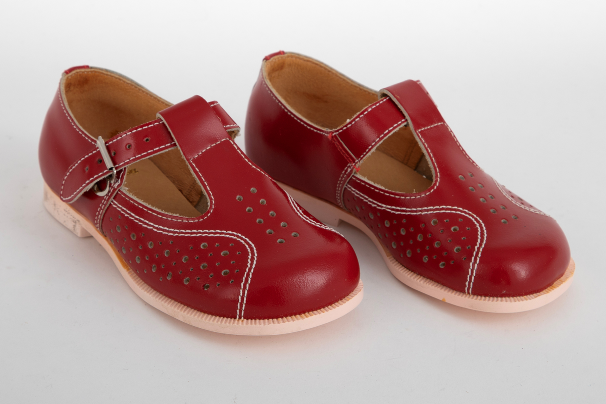 Barnesko i rødt skinn med gjennomhullete mønsterpartier og hvite stikninger. Pensko med vristrem gjennom stropp midt foran og spenne i sidene.