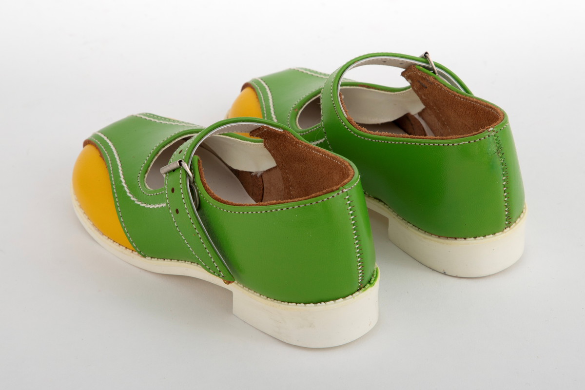 Barnesko i gressgrønt lær med gule detaljer på hver side av forstykket. Hvite stikninger og vristrem med spenne. Skoene har hvite såler i syntetisk gummi.