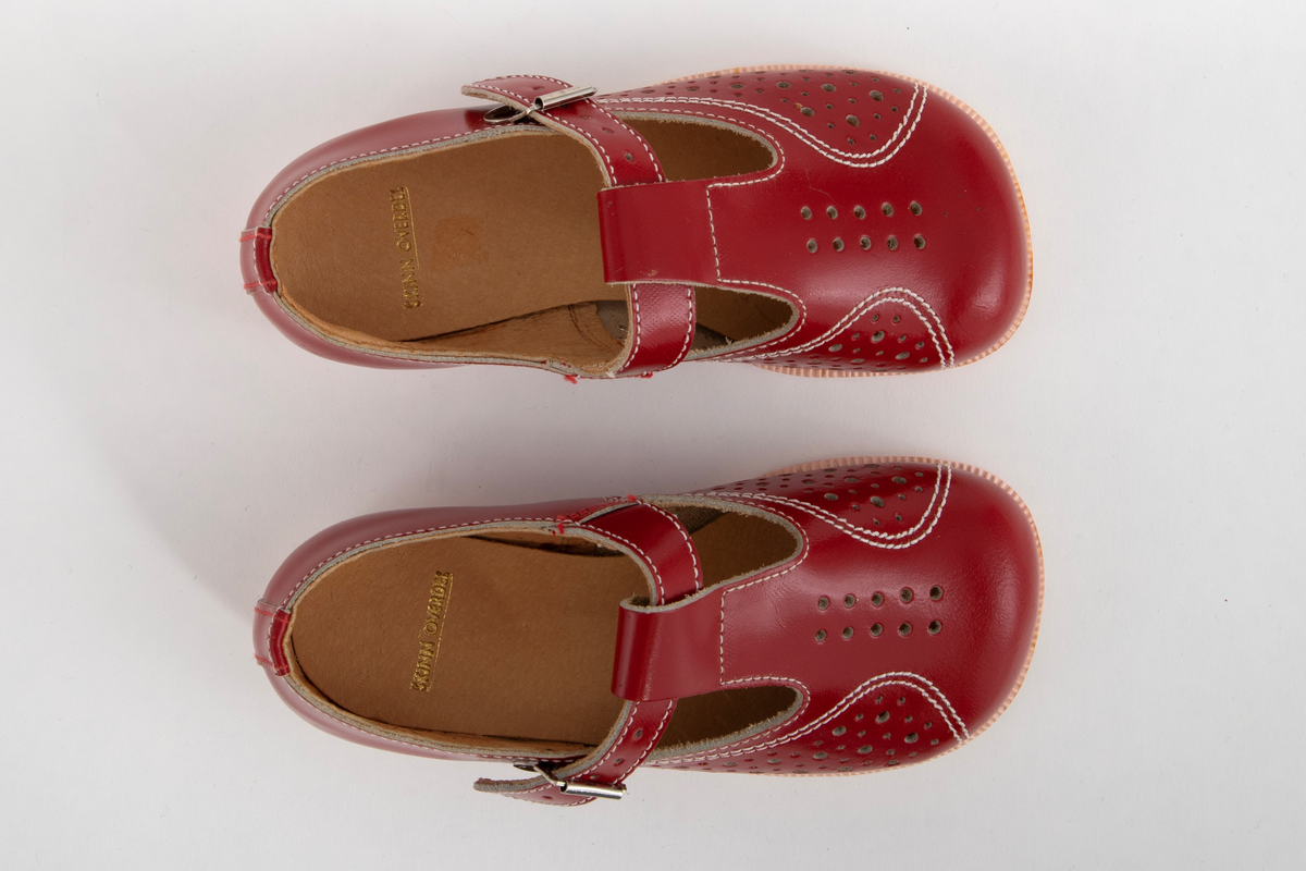 Barnesko i rødt skinn med gjennomhullete mønsterpartier og hvite stikninger. Pensko med vristrem gjennom stropp midt foran og spenne i sidene.