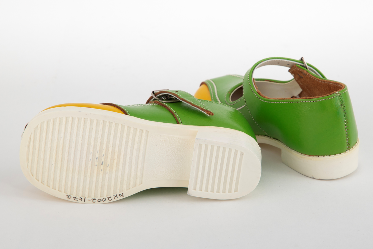 Barnesko i gressgrønt lær med gule detaljer på hver side av forstykket. Hvite stikninger og vristrem med spenne. Skoene har hvite såler i syntetisk gummi.