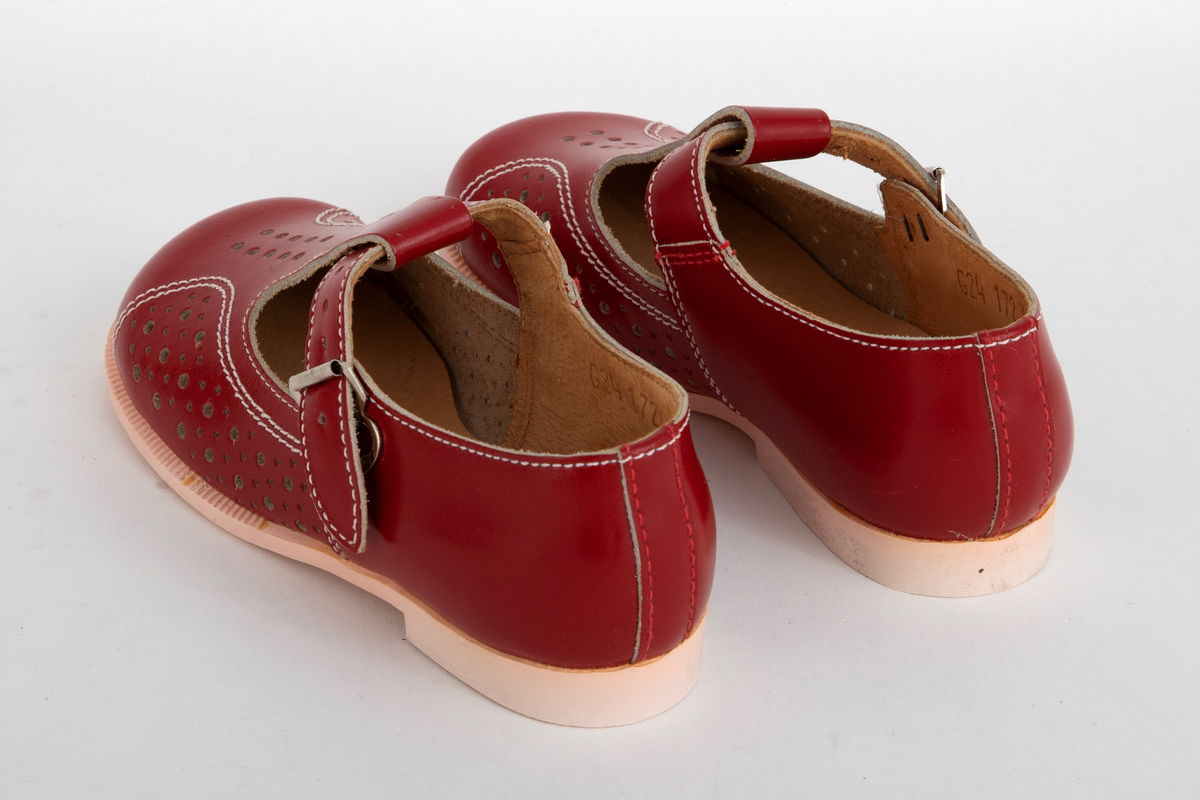 Barnesko i rødt skinn med gjennomhullete mønsterpartier og hvite stikninger. Pensko med vristrem gjennom stropp midt foran og spenne i sidene.