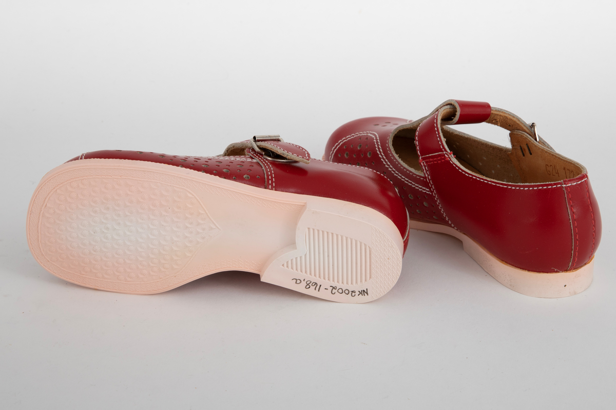 Barnesko i rødt skinn med gjennomhullete mønsterpartier og hvite stikninger. Pensko med vristrem gjennom stropp midt foran og spenne i sidene.