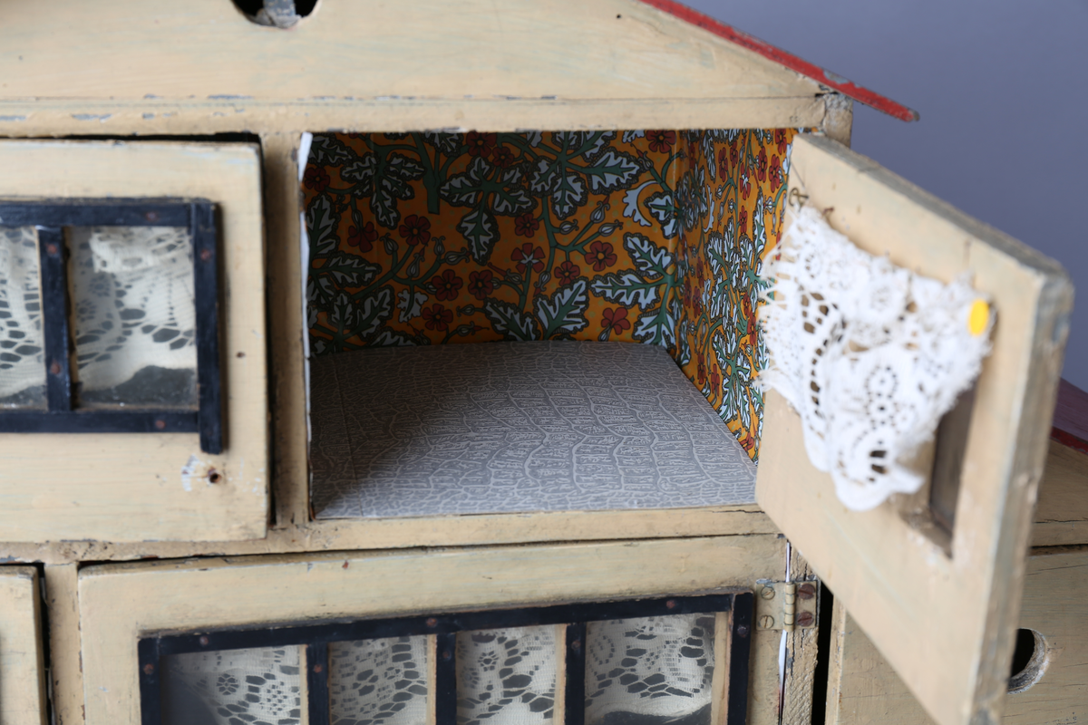 Rektangulært dukkehus i treverk med tre vegger, festet tak og dører i front. Huset er gult på utsiden og har tre etasjer, hvor loftet er lukket, men har et mindre hull i front. Taket er rødt. Dørene har vinduer med brunt listverk og hvite blondegardiner på innsiden. Tre gulvflater er belagt med grå linoleum, to gulvflater mangler belegg. Fire av rommene har tapet på veggene, alle i rikt detaljerte mønstre i flere farger. Taket i andre etasje har et hvitt intrikat mønster. Tomme rom uten særlige tegn til bruksområder.