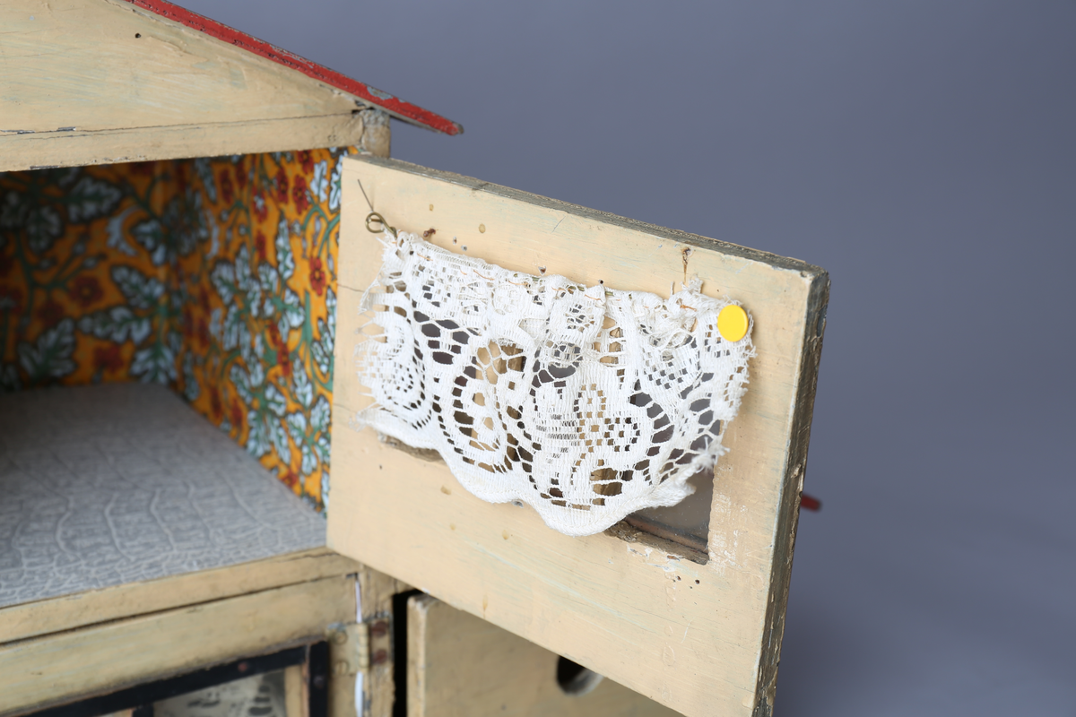 Rektangulært dukkehus i treverk med tre vegger, festet tak og dører i front. Huset er gult på utsiden og har tre etasjer, hvor loftet er lukket, men har et mindre hull i front. Taket er rødt. Dørene har vinduer med brunt listverk og hvite blondegardiner på innsiden. Tre gulvflater er belagt med grå linoleum, to gulvflater mangler belegg. Fire av rommene har tapet på veggene, alle i rikt detaljerte mønstre i flere farger. Taket i andre etasje har et hvitt intrikat mønster. Tomme rom uten særlige tegn til bruksområder.