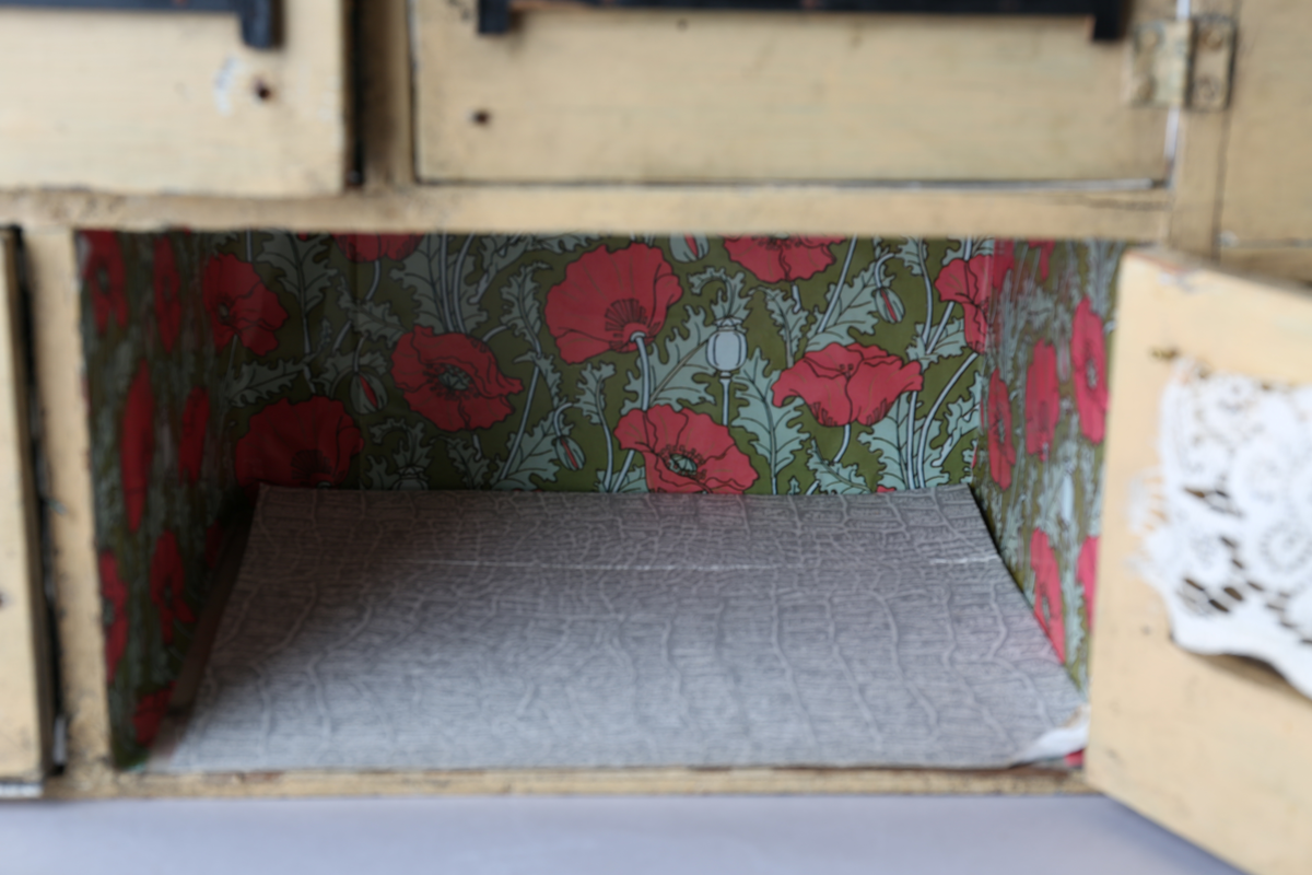 Rektangulært dukkehus i treverk med tre vegger, festet tak og dører i front. Huset er gult på utsiden og har tre etasjer, hvor loftet er lukket, men har et mindre hull i front. Taket er rødt. Dørene har vinduer med brunt listverk og hvite blondegardiner på innsiden. Tre gulvflater er belagt med grå linoleum, to gulvflater mangler belegg. Fire av rommene har tapet på veggene, alle i rikt detaljerte mønstre i flere farger. Taket i andre etasje har et hvitt intrikat mønster. Tomme rom uten særlige tegn til bruksområder.