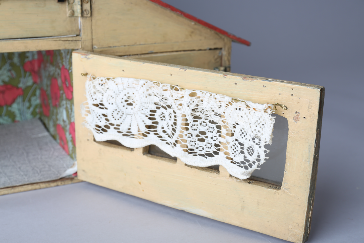 Rektangulært dukkehus i treverk med tre vegger, festet tak og dører i front. Huset er gult på utsiden og har tre etasjer, hvor loftet er lukket, men har et mindre hull i front. Taket er rødt. Dørene har vinduer med brunt listverk og hvite blondegardiner på innsiden. Tre gulvflater er belagt med grå linoleum, to gulvflater mangler belegg. Fire av rommene har tapet på veggene, alle i rikt detaljerte mønstre i flere farger. Taket i andre etasje har et hvitt intrikat mønster. Tomme rom uten særlige tegn til bruksområder.