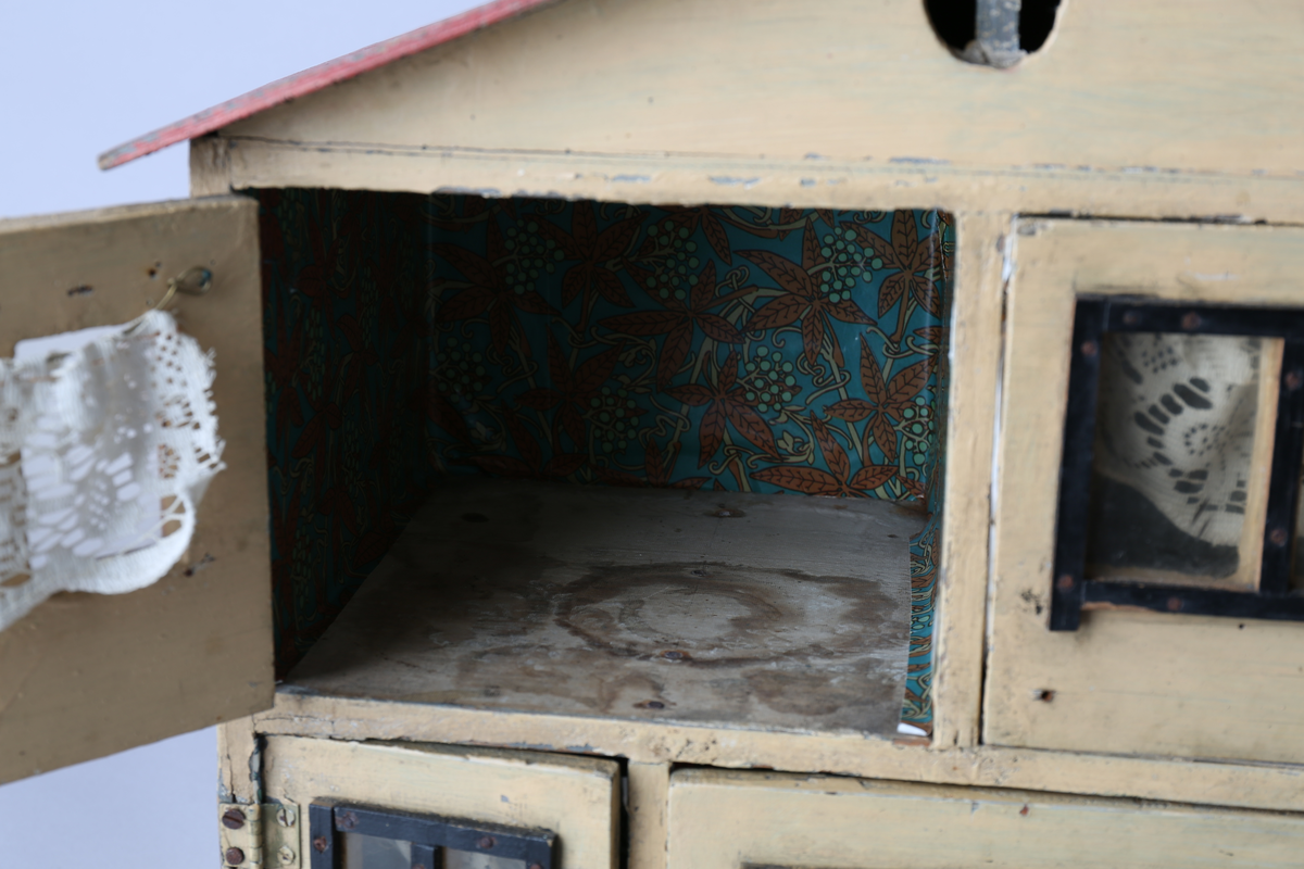 Rektangulært dukkehus i treverk med tre vegger, festet tak og dører i front. Huset er gult på utsiden og har tre etasjer, hvor loftet er lukket, men har et mindre hull i front. Taket er rødt. Dørene har vinduer med brunt listverk og hvite blondegardiner på innsiden. Tre gulvflater er belagt med grå linoleum, to gulvflater mangler belegg. Fire av rommene har tapet på veggene, alle i rikt detaljerte mønstre i flere farger. Taket i andre etasje har et hvitt intrikat mønster. Tomme rom uten særlige tegn til bruksområder.