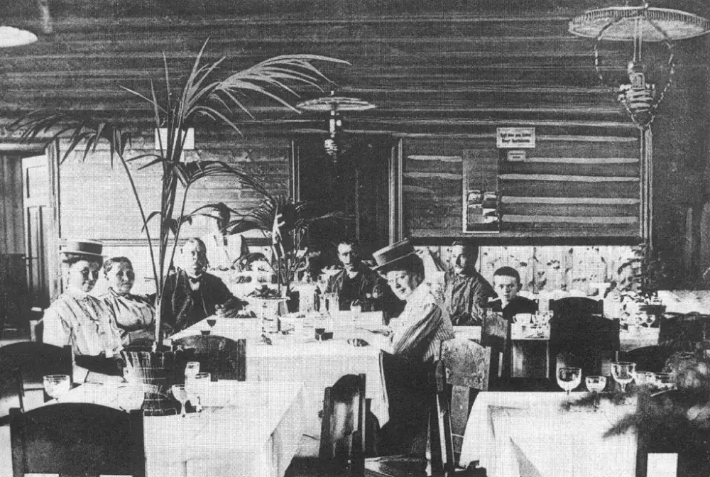 Restauranten på Krøderen stasjon. En gruppe på sju ser mot kameraet. De er pent kledde. Den yngste er et barn, og den eldste er en eldre mann. En servitør står i bakgrunnen. Det er hvite duker og eksotiske planter på bordene.