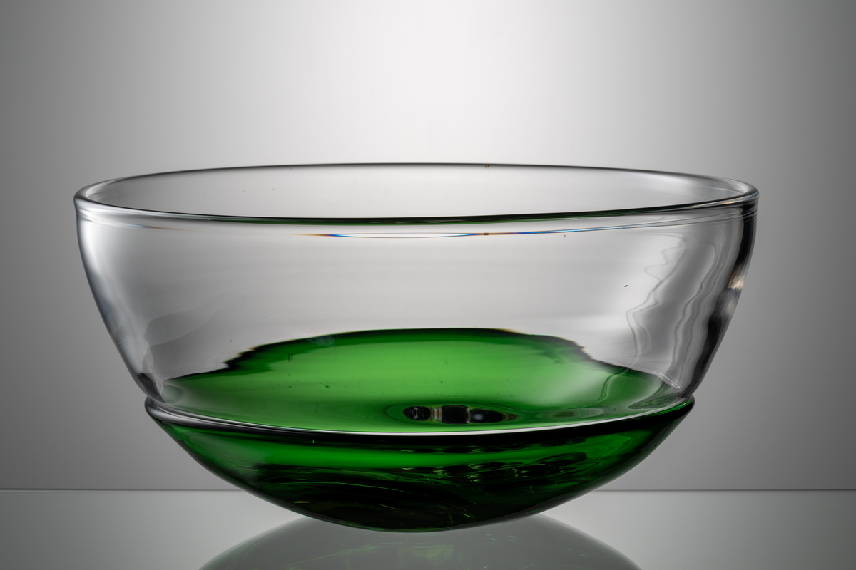 Formgivare: Lars Hellsten. Skål med närmast halvsfärisk form. I nedre delen en driven grön skål med dekor i form av ”medaljonghalvor” med abstrakt mönster. Över den en nedtryckt ofärgad skål. Sammanfogade varmt i hyttan.