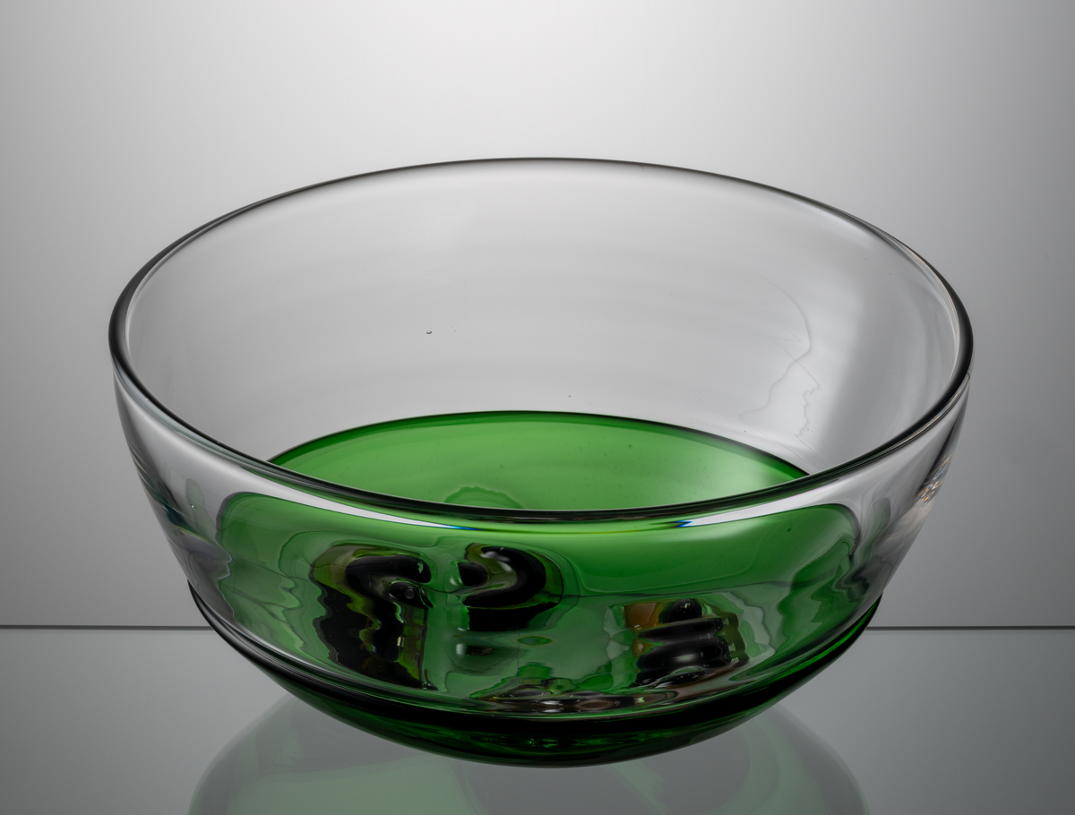 Formgivare: Lars Hellsten. Skål med närmast halvsfärisk form. I nedre delen en driven grön skål med dekor i form av ”medaljonghalvor” med abstrakt mönster. Över den en nedtryckt ofärgad skål. Sammanfogade varmt i hyttan.