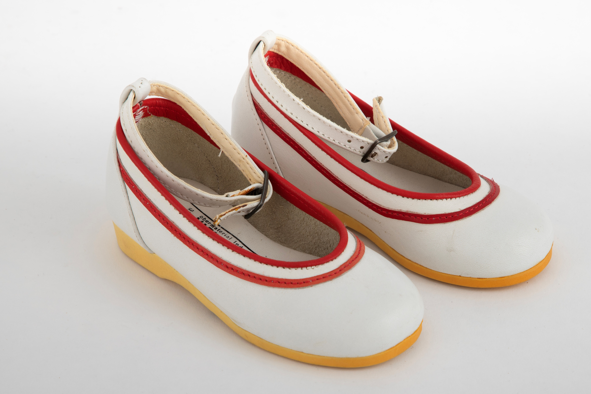 Barnesko i hvitt skinn med røde striper langs åpningen. Hvit ankelrem med metallspenne. Skoene har lys såle, som har gulnet.