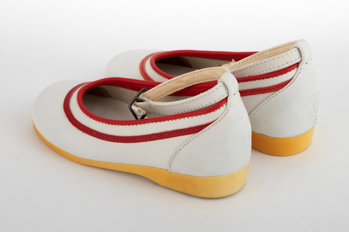 Barnesko i hvitt skinn med røde striper langs åpningen. Hvit ankelrem med metallspenne. Skoene har lys såle, som har gulnet.