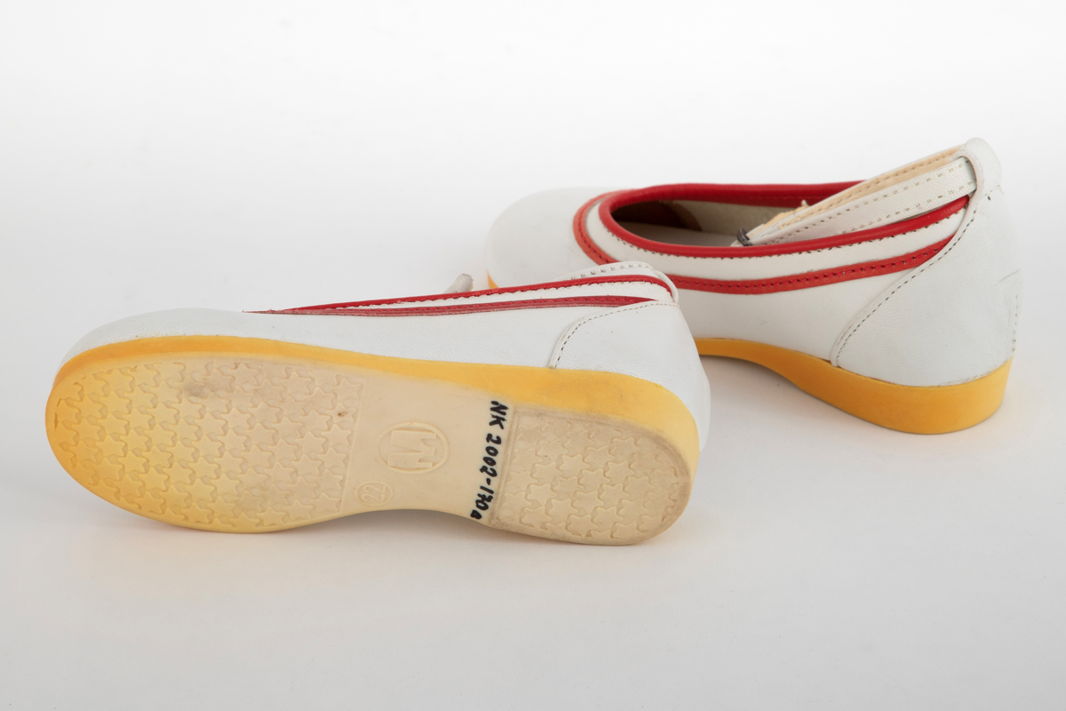 Barnesko i hvitt skinn med røde striper langs åpningen. Hvit ankelrem med metallspenne. Skoene har lys såle, som har gulnet.
